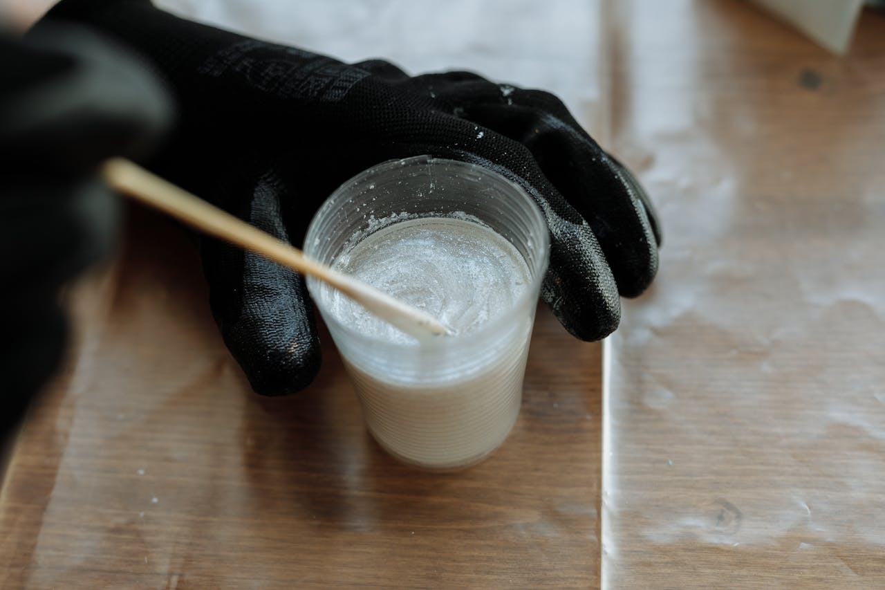 about-us Detailed view of hands with gloves mixing resin in a plastic cup with a stirrer on a wooden surface.